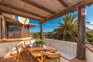Une table en bois avec des chaises et un banc, une antenne parabolique, et une vue sur des palmiers et des collines.