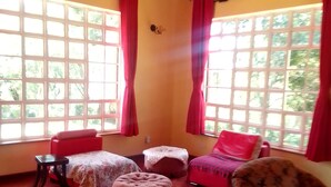 A room with wooden flooring, a wooden ceiling, and large windows with red curtains. There are two red sofas, a floral armchair, and a small table. - TIGONI GEM GARDENS (Limuru)