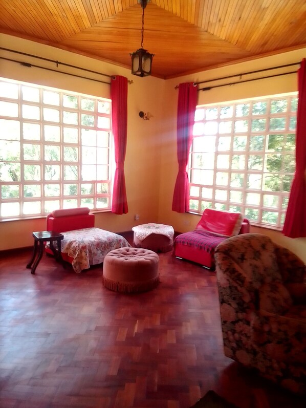 A room with wooden flooring, a wooden ceiling, and large windows with red curtains. There are two red sofas, a floral armchair, and a small table.