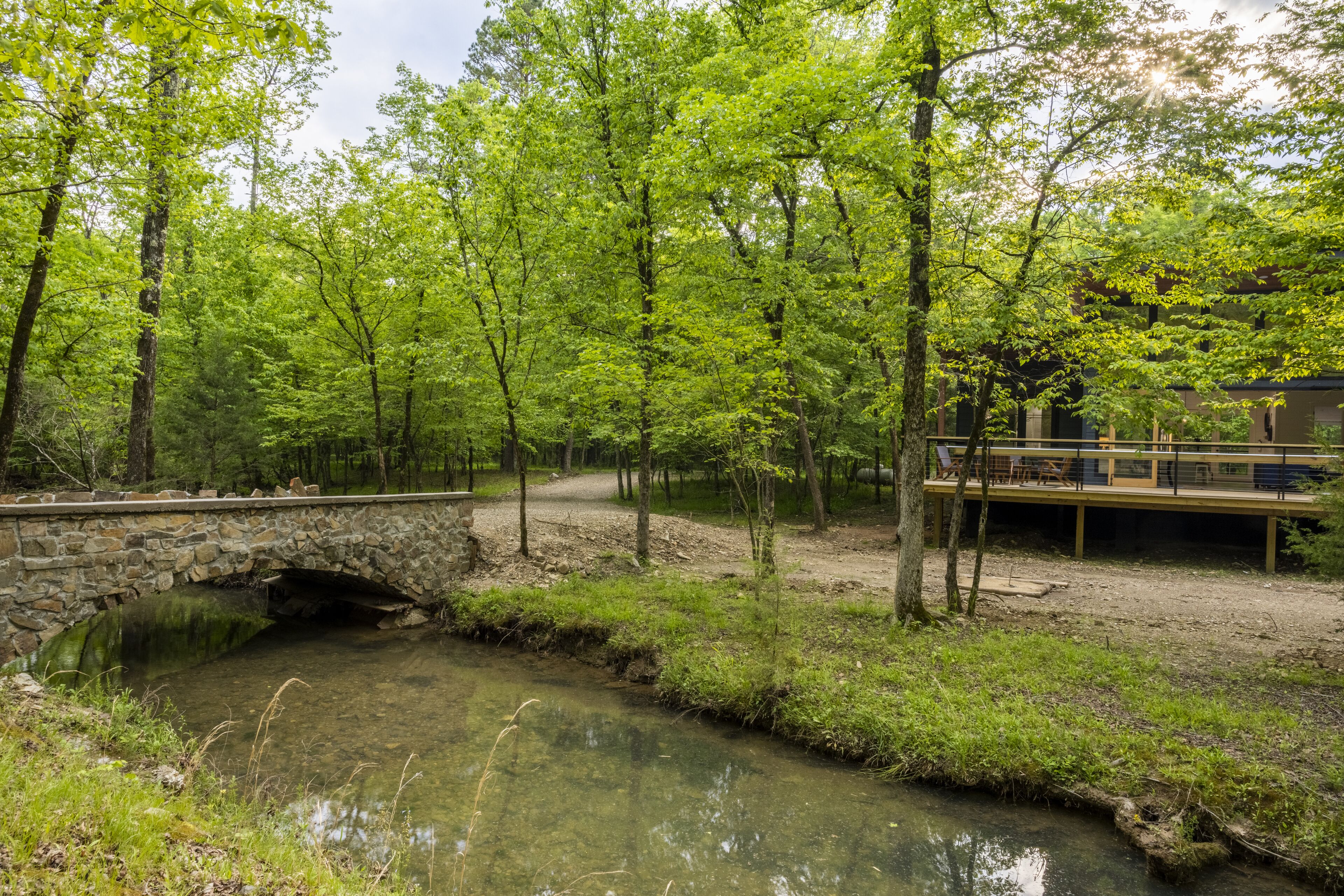 Un pont de pierre enjambe un ruisseau aux eaux claires, dans une zone boisée, et est doté d’une terrasse en bois ainsi que de bancs.