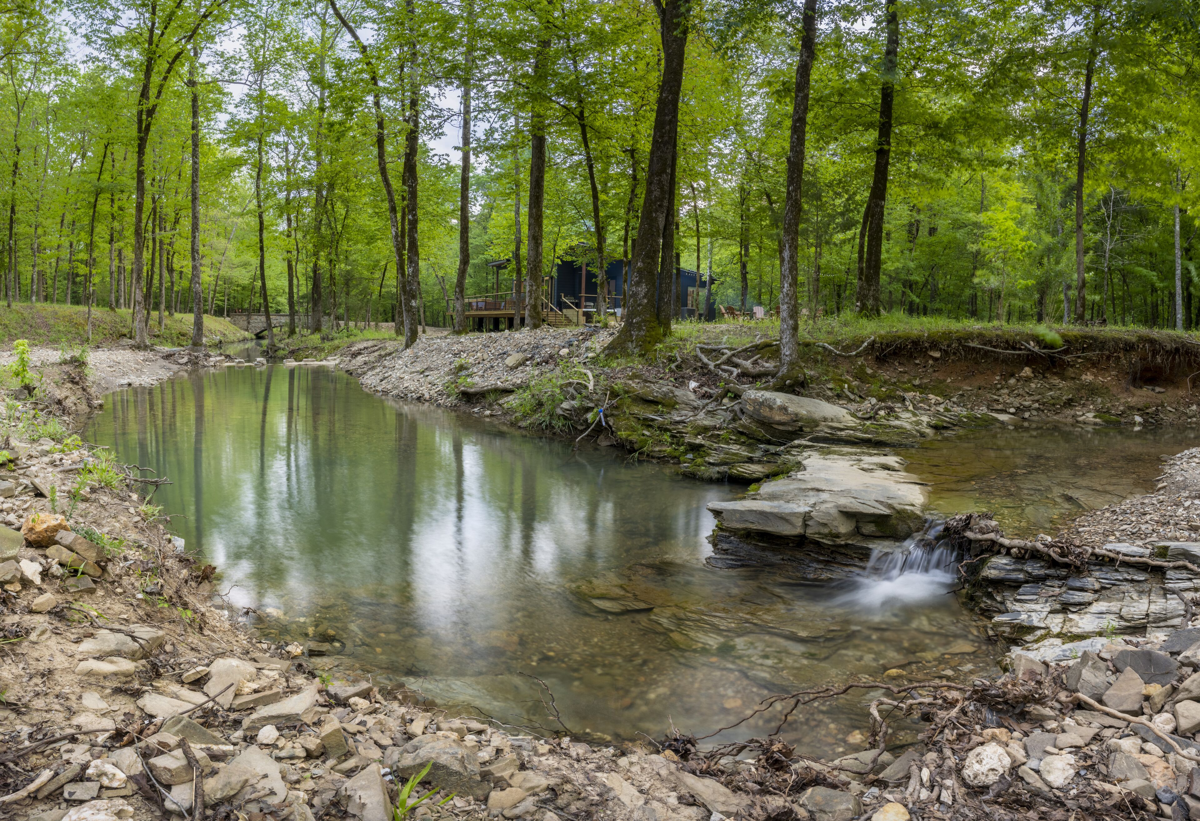 A natural outdoor setting with a small stream, surrounded by trees and a wooden structure.