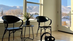 A room with two black chairs, a small table with a plant, and a remote control on a white table. Large windows offer a view of snow-covered mountains and a clear sky. - Paradise Valley View Getaway (Livingston)