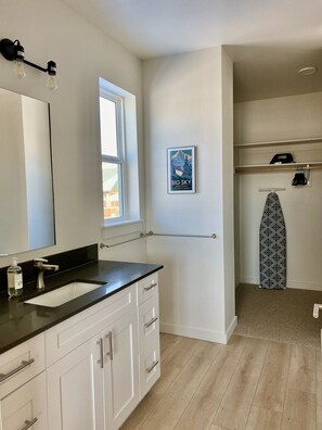 A modern bathroom with a large mirror, a black countertop, a white sink, and a window. There is a poster on the wall and a surfboard on a shelf.
