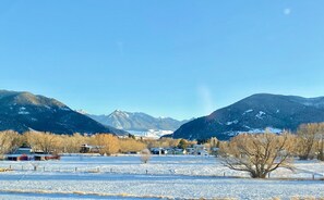 A rural landscape with snow-covered fields, a few houses, and mountains in the background.