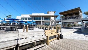A wooden deck with outdoor seating and umbrellas, a building with large windows and a balcony, and string lights overhead. - Excellent Location, Beautiful House Sleeps 10 and Will Feel Like Home!  (Orange Beach)