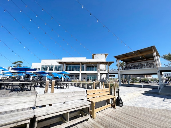A wooden deck with outdoor seating and umbrellas, a building with large windows and a balcony, and string lights overhead.