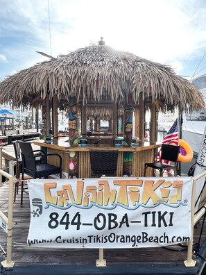 A tiki bar with a thatched roof, wooden tables, and chairs. A banner displays the name and contact information.