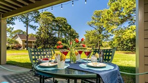 Um pátio externo com mesa redonda para dois, com toalha de mesa azul, duas cadeiras, um vaso com tulipas vermelhas e um jarro com bebidas geladas.