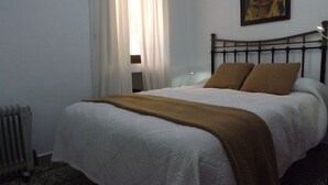 A neatly made bed with a white duvet and two brown pillows, a radiator, and a window with white curtains. - Casa rural rodeada de arboles frutales con piscinas privadas y wifi (Cómpeta)