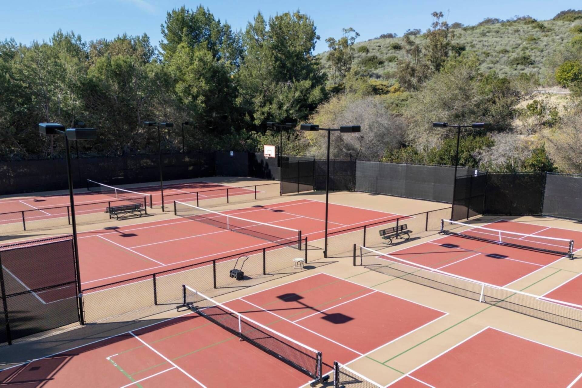 Una cancha de tenis exterior con varias canchas, bancos e iluminarias.