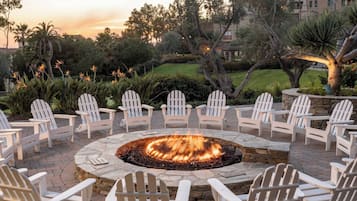 A circular outdoor seating area with a fire pit surrounded by white Adirondack chairs, set on a stone patio with a view of a landscaped garden and buildings in the background.