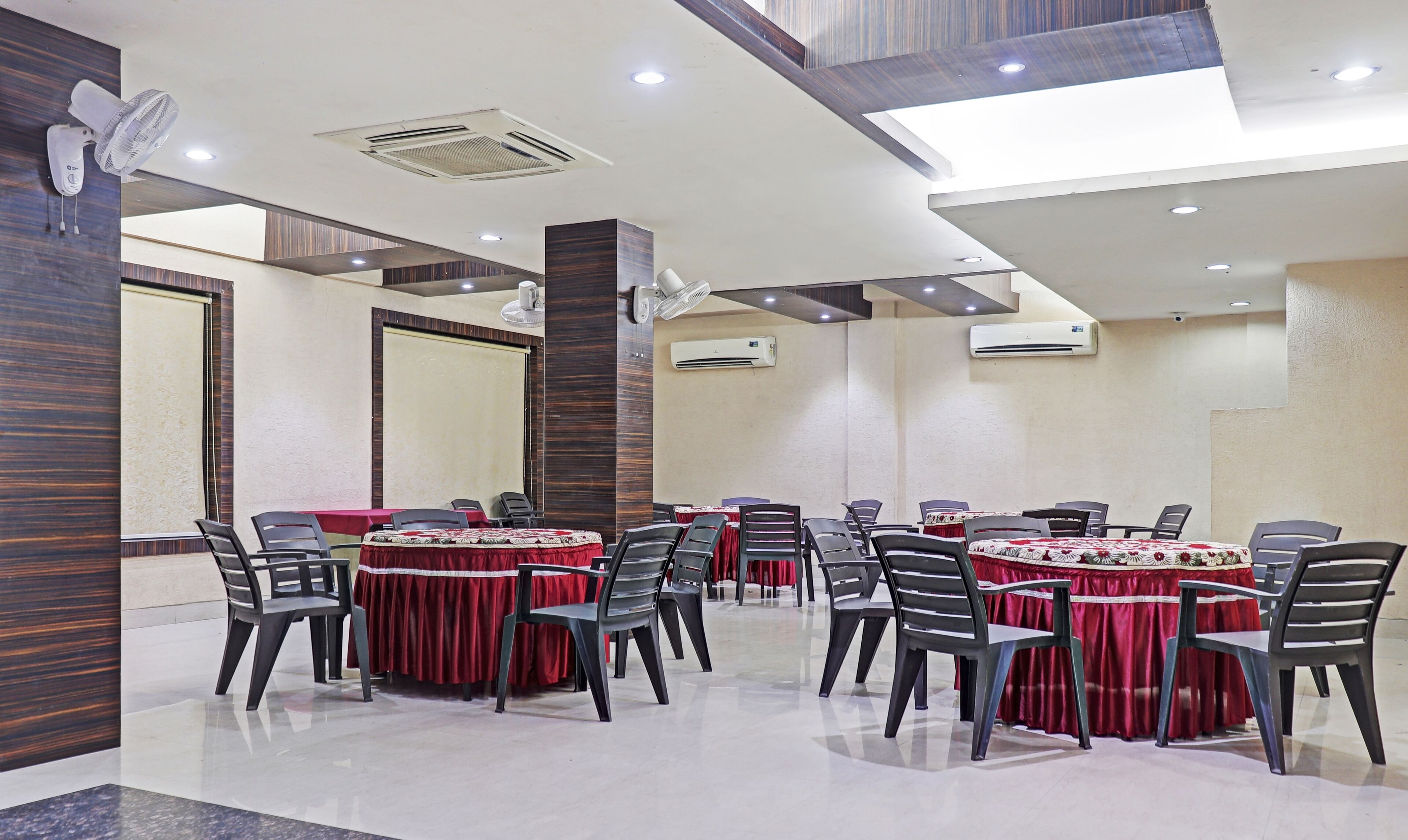 A conference room with round tables, chairs, and red tablecloths. There are air conditioning units on the walls and ceiling fans.