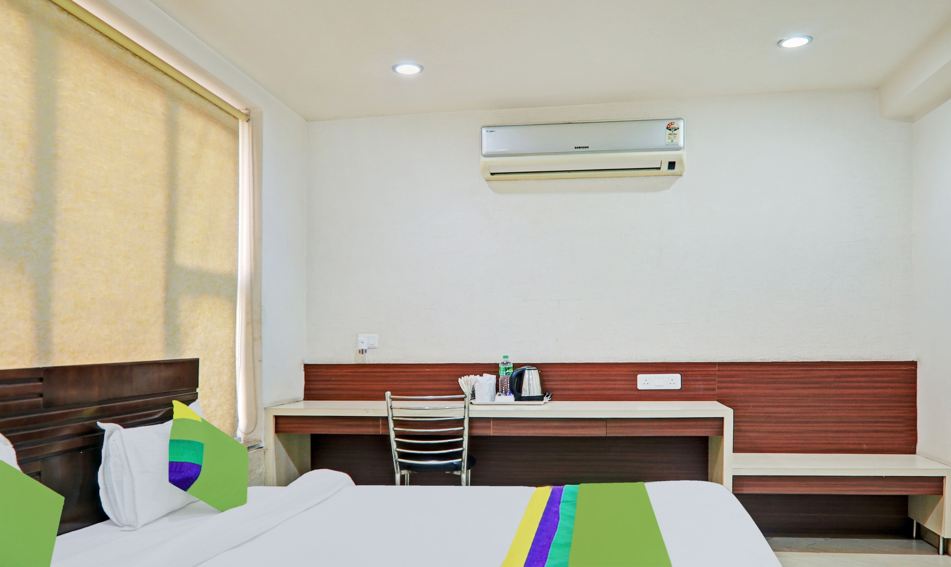 A hotel room with a bed, a desk, a chair, an air conditioning unit, and a window with curtains.