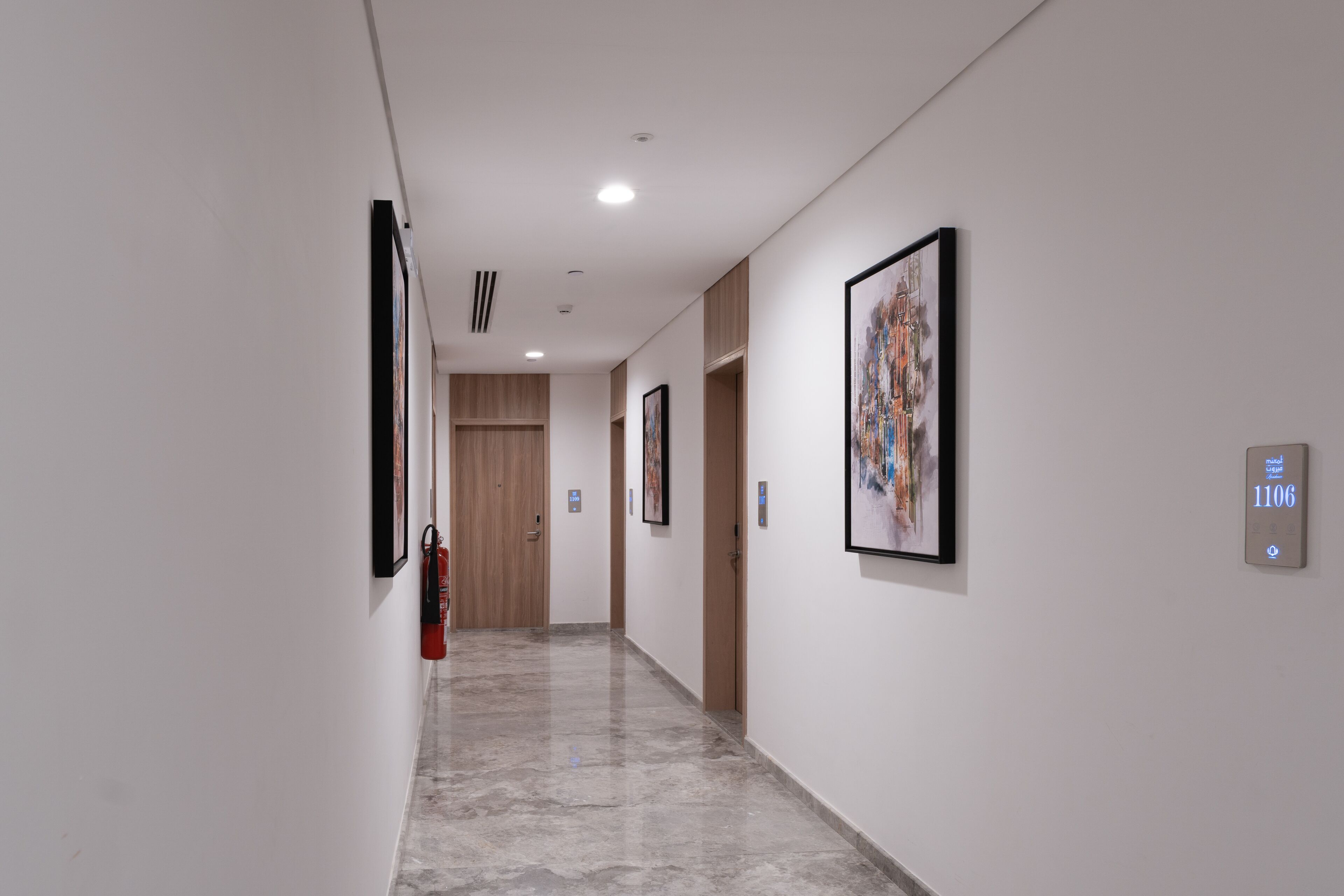 A hallway with framed artwork on the walls, a fire extinguisher mounted on the wall, and a digital display showing room number 1106.