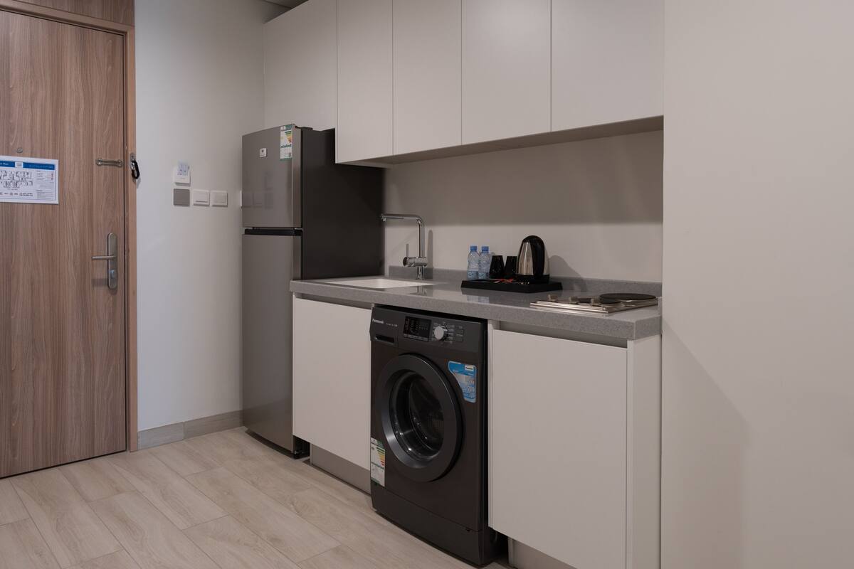 Une cuisine compacte équipée d’un lave-linge, d’un réfrigérateur et d’un plan de travail sur lequel se trouvent une machine à café et des bouteilles d’eau.