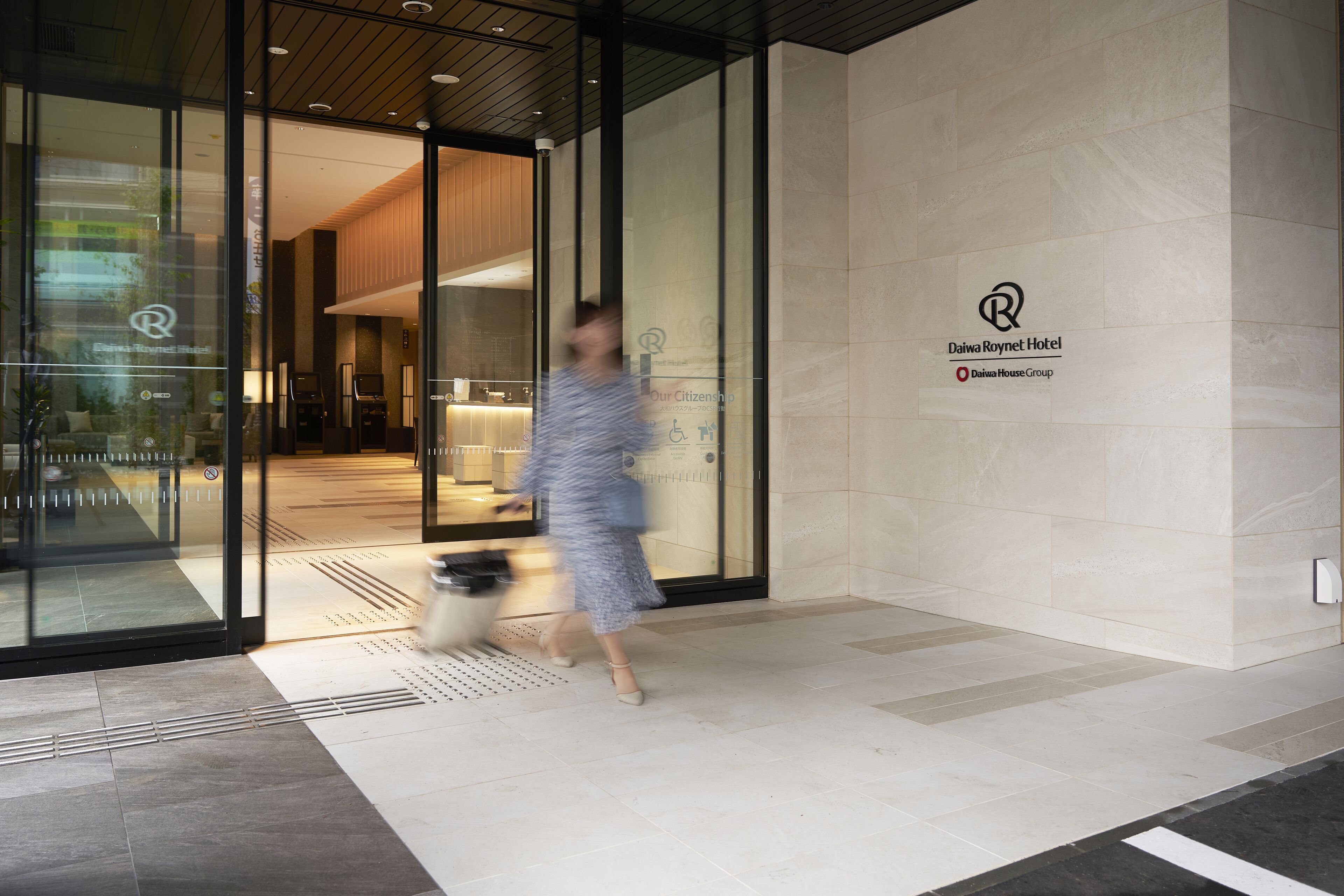 A modern hotel entrance with a glass facade, a visible interior with seating, and a logo for Daiwa Roynet Hotel.