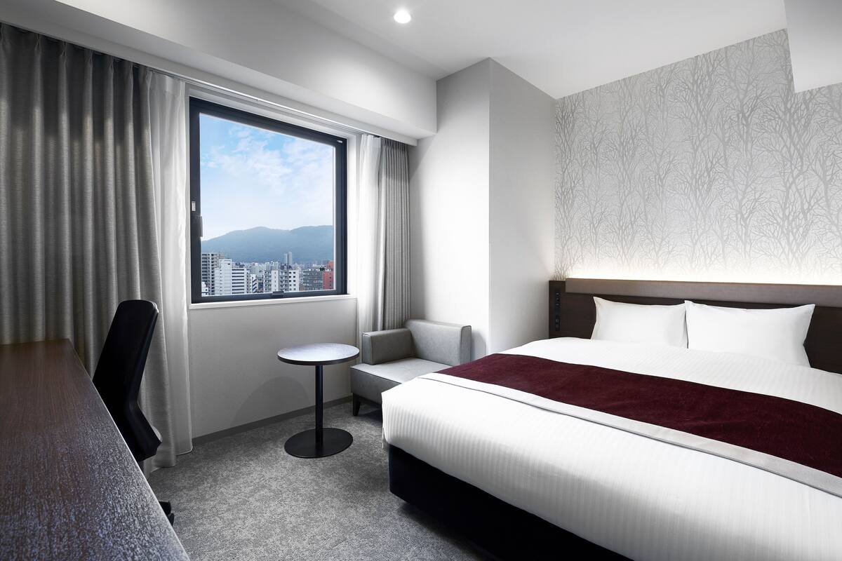 Une chambre d’hôtel avec un lit, un bureau, une chaise, une petite table et une fenêtre donnant sur la ville.