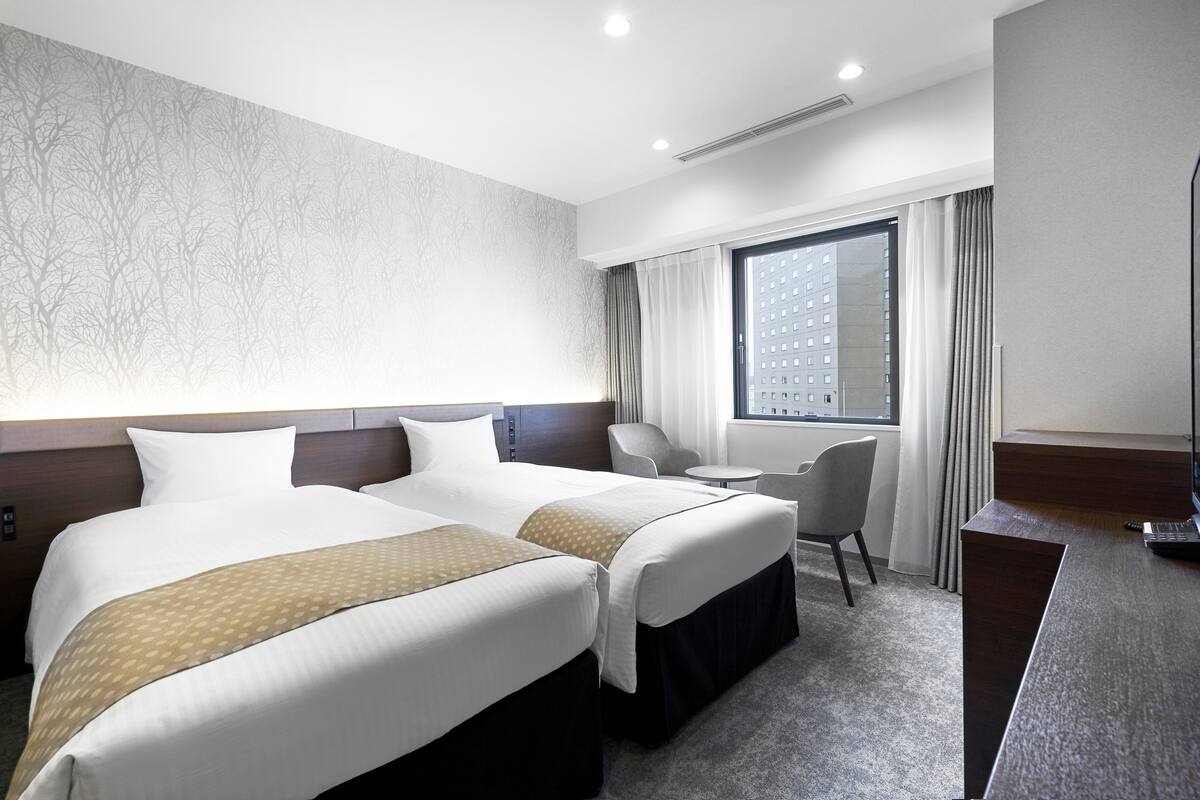 Une chambre d’hôtel avec deux lits, un bureau, une chaise et une grande fenêtre avec des rideaux.