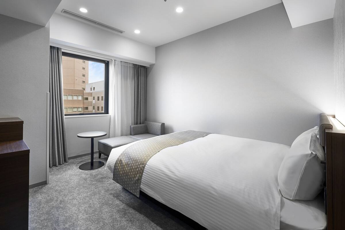 Une chambre d’hôtel avec un lit, une petite table, une chaise et une fenêtre avec des rideaux.
