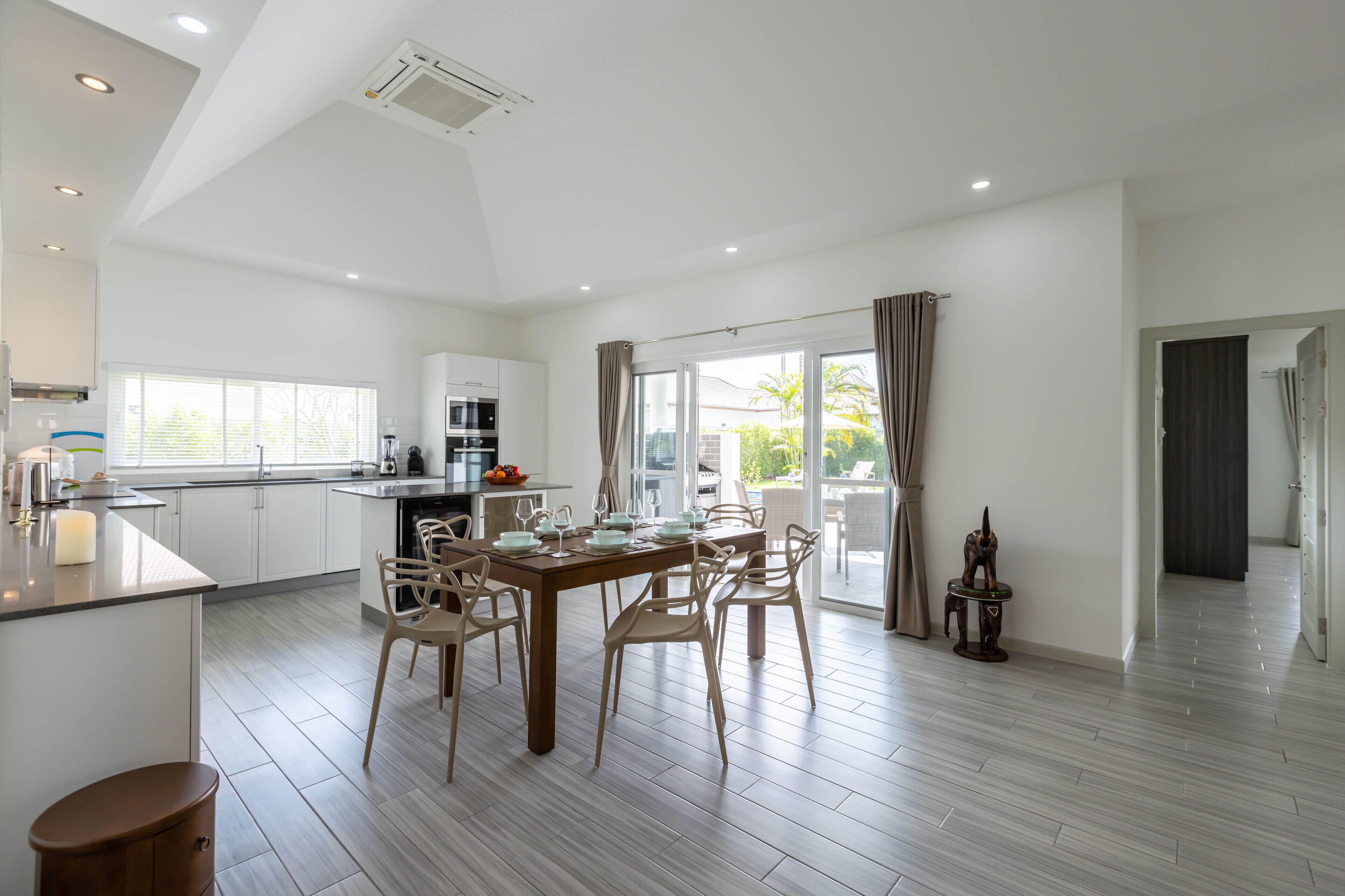 A modern kitchen with white cabinets, a dining area with a wooden table and chairs, and a large window with curtains.