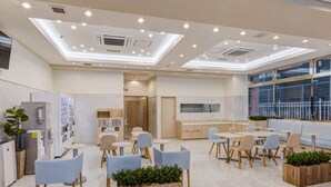 Espaço interior moderno com design limpo e minimalista, apresentando uma área de estar central com mesas e cadeiras, rodeada por estantes e uma cozinha.
