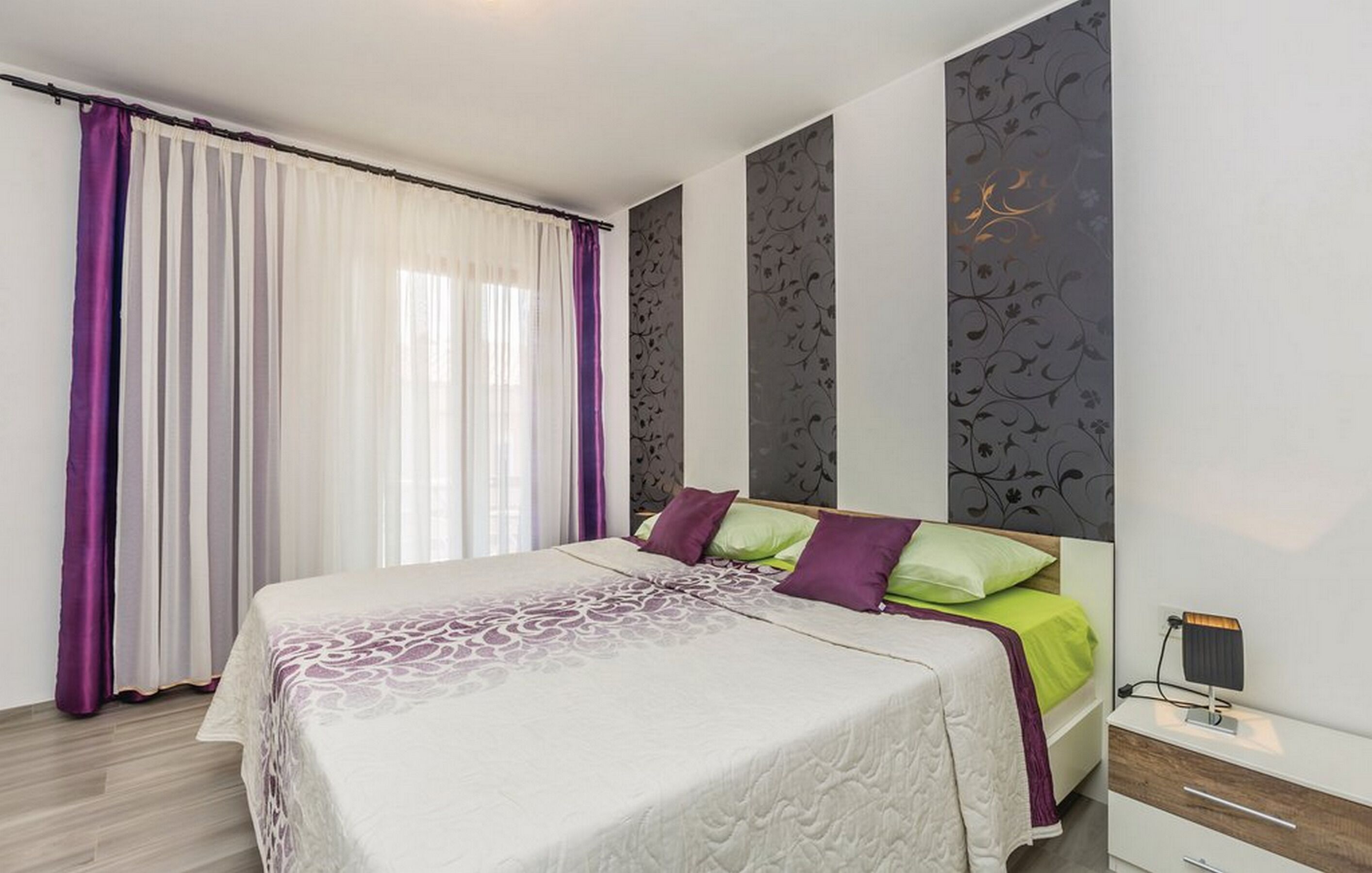 Een moderne slaapkamer met een groot bed, paarse en groene kussens, een nachtkastje met een lamp en een raam met dunne gordijnen.
