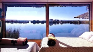 A room with a large window overlooking a calm body of water, a bed with a rolled-up towel, and a small table with a bottle. - Room-suma Jakana Habitación Vista Al Ama (Puno)