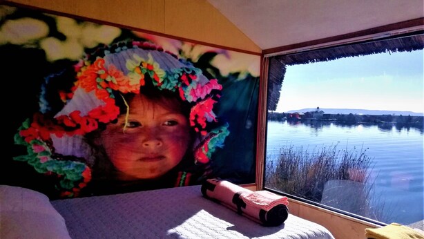 A bedroom with a bed, pillows, a window with a view of a lake, and a colorful wall decoration. - Room-suma Jakana Habitación Vista Al Ama (Puno)