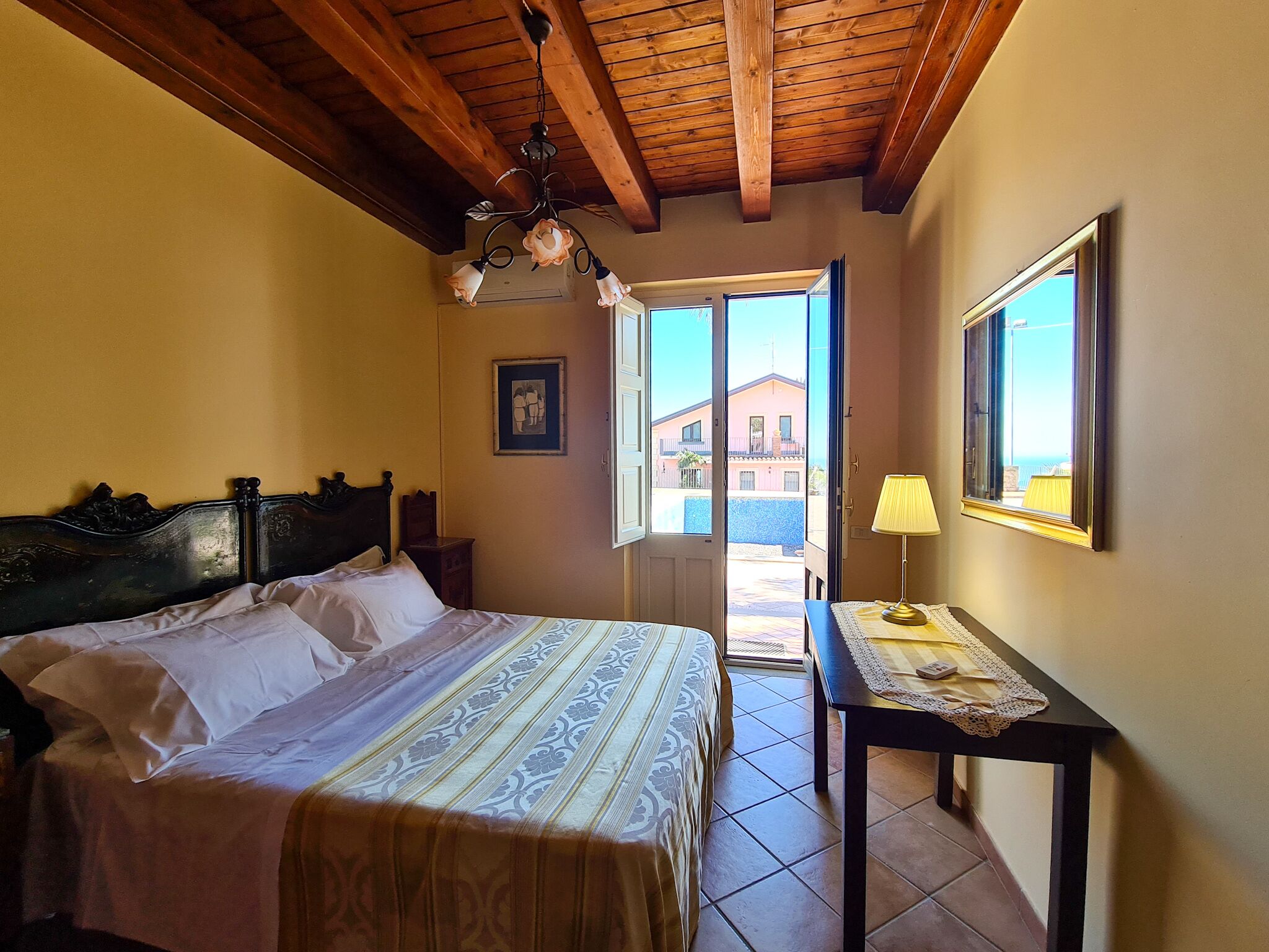 A bedroom with a bed, a nightstand, a table with a lamp, a framed picture, and a view of the outside.