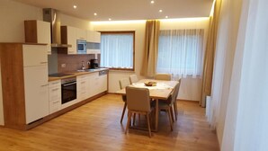 Eine kleine Küche mit modernen Geräten, ein Essbereich mit Tisch und Stühlen sowie große Fenster mit durchsichtigen Vorhängen.