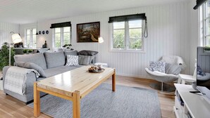 Amplia sala de estar con una mesa de centro de madera, un sofá gris y un sillón blanco. Hay un cuadro en la pared, una ventana con vistas a la vegetación y un televisor sobre un mueble blanco.