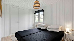 Un dormitorio con una cama grande, una ventana con cortina negra, una lámpara colgante de mimbre y paredes blancas con paneles.