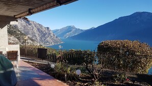 Eine Terrasse mit Blick auf einen See und Berge, mit Metallgeländer, Blumentöpfen und Gartenmöbeln.