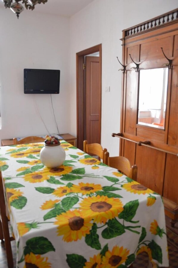 Un coin repas avec une table recouverte d’une nappe à motifs de tournesols, un vase rempli de fruits et un téléviseur fixé au mur.
