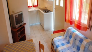 A compact room with a bed, a small table, a television, a kitchenette, and a window with curtains. - Villa in Lignano Pineta Close to the Beach (Lignano Sabbiadoro)