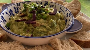 Un tazón de guacamole con cebollines y pimientos rojos, rodeado de rebanadas de pan en un plato.