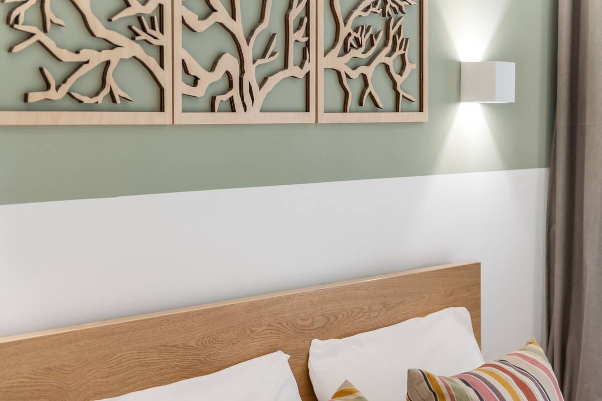 A neatly made bed with white bedding and striped pillows, a wooden headboard, and a wall-mounted light fixture.