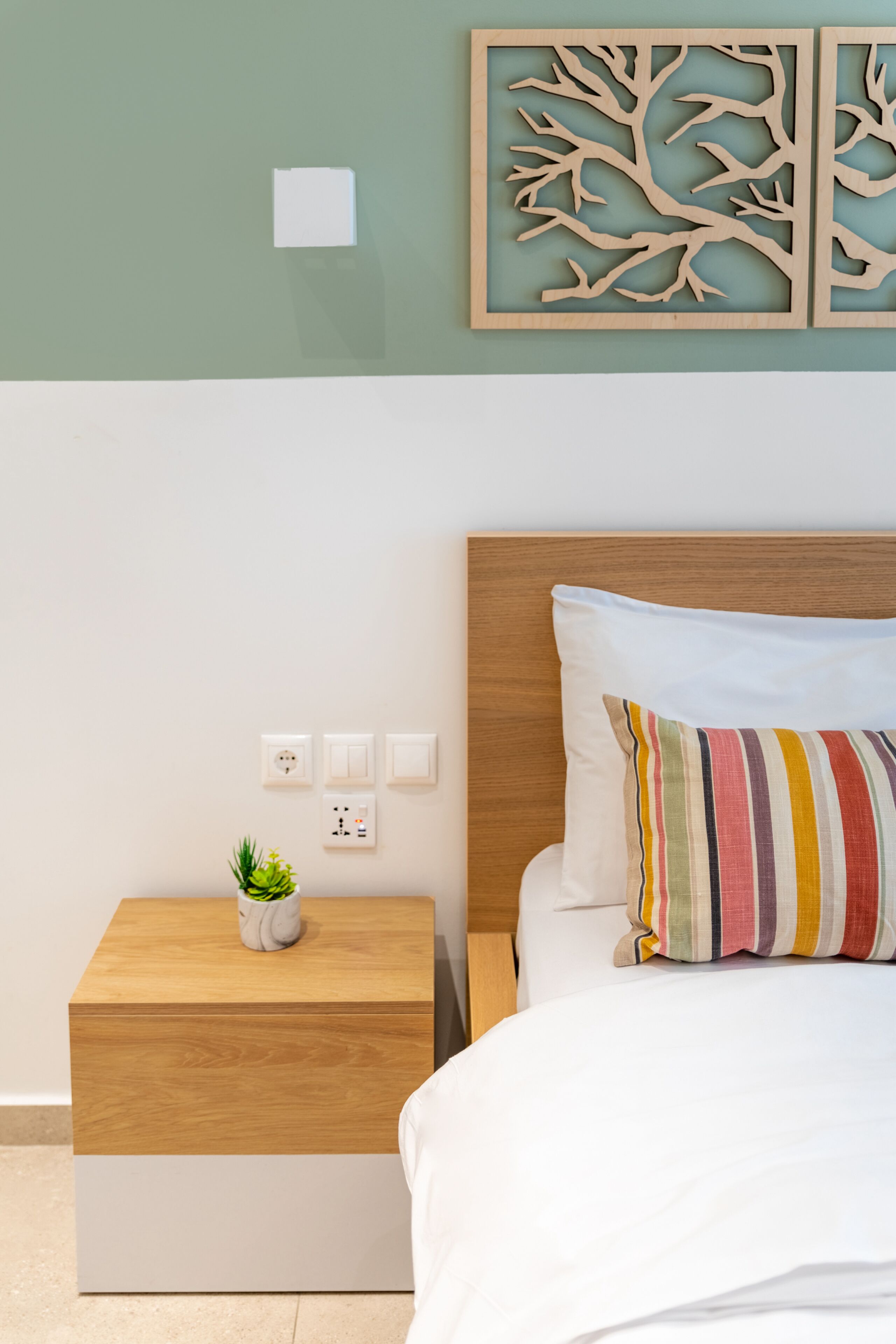 A neatly arranged bedroom with a wooden nightstand, a small potted plant, a bed with a striped pillow, and a wall-mounted power outlet.
