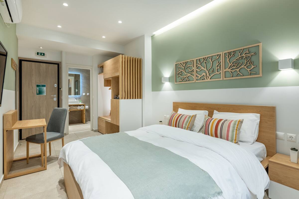 A modern hotel room with a bed, a desk, and a chair. There is a wooden wardrobe and a mirror.