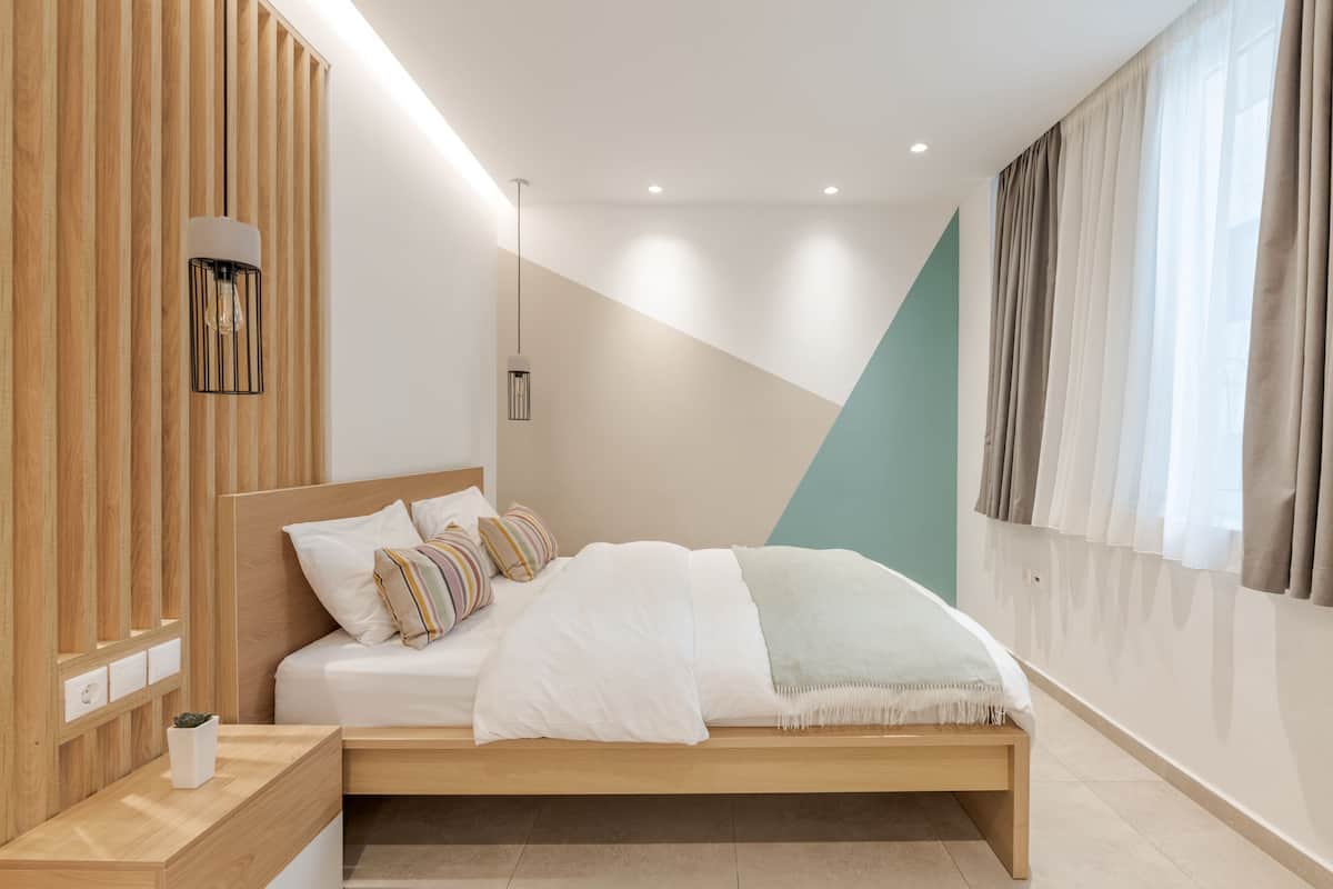 A modern bedroom with a wooden headboard, a bedside table with a plant, and a bed with white bedding and a light blue throw.