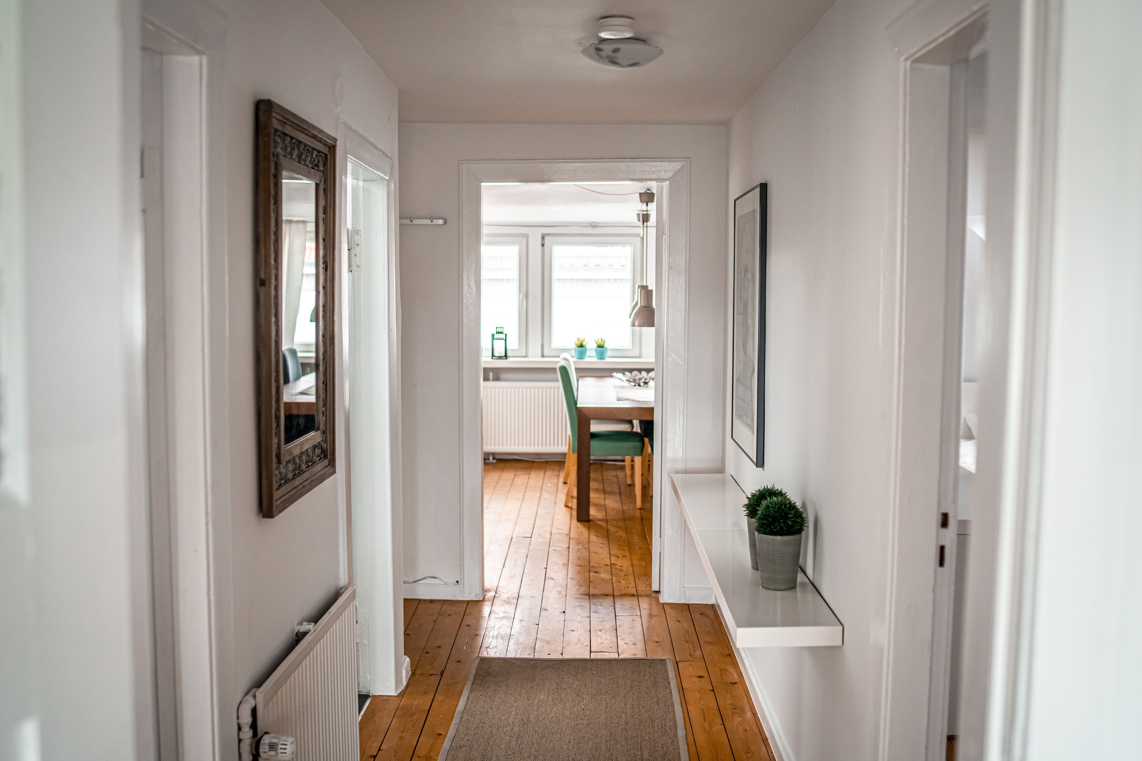 A hallway with a wooden floor, white walls, and a door leading to a dining area with a table and chairs.