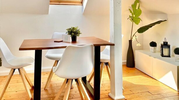 Quarto moderno estilo loft com teto inclinado, mesa de jantar de madeira, cadeiras brancas, uma janela grande e uma planta.