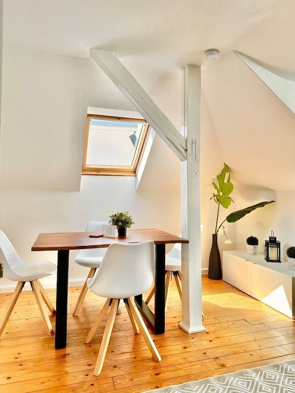 A modern loft bedroom with a sloped ceiling, a wooden dining table, white chairs, a large window, and a plant.