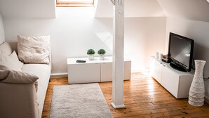 Uma sala de estar moderna e minimalista com piso de madeira, móveis brancos, uma televisão e uma pequena planta em cima de um console branco.