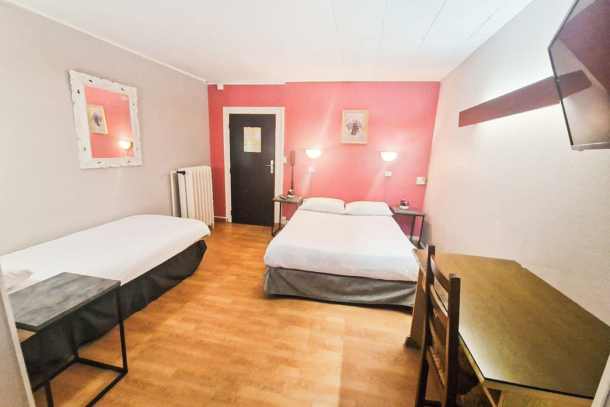 A room with two single beds, a small table, and a chair. There is a radiator, a door, and a wall-mounted light fixture.