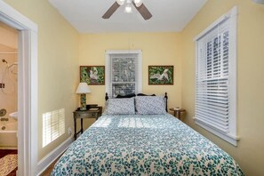 Quarto com cama, ventilador de teto, janela com persianas e quadros nas paredes.