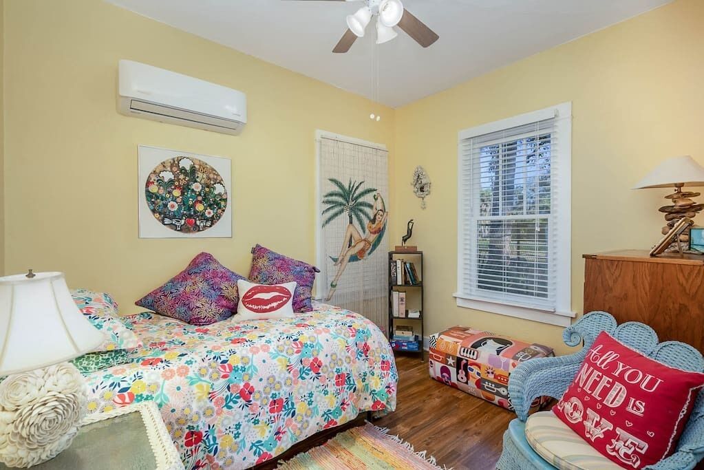 A bedroom with a bed, a nightstand, a lamp, a window with blinds, a ceiling fan, and a wall-mounted air conditioner.