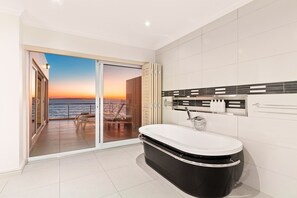 Une salle de bain moderne dotée d’une grande baignoire et d’une porte en verre donnant sur un balcon d’où l’on peut admirer le coucher de soleil sur la mer.