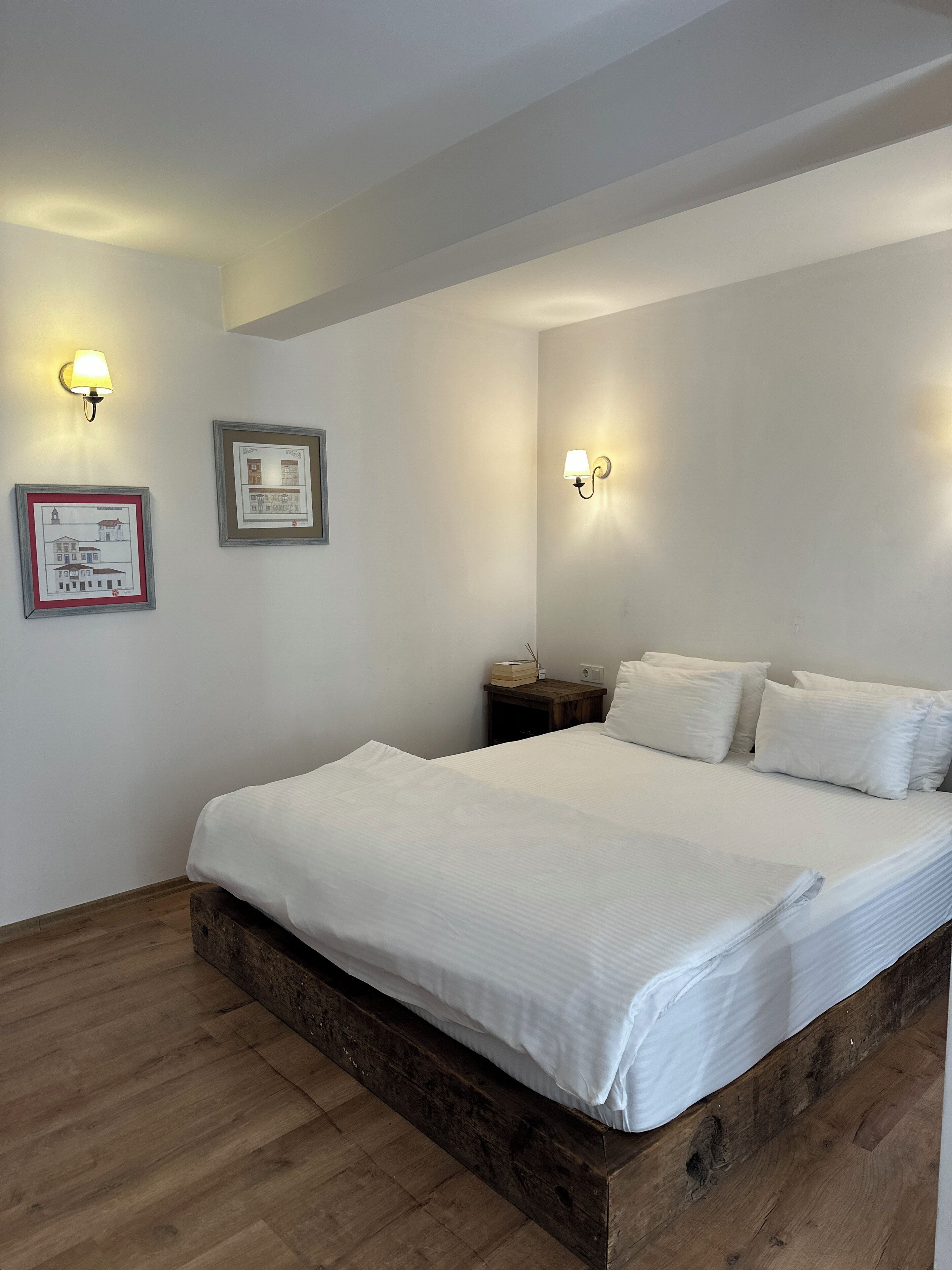 Quarto com estrutura de cama de madeira, roupa de cama branca, duas luminárias fixadas na parede e desenhos arquitetônicos emoldurados.