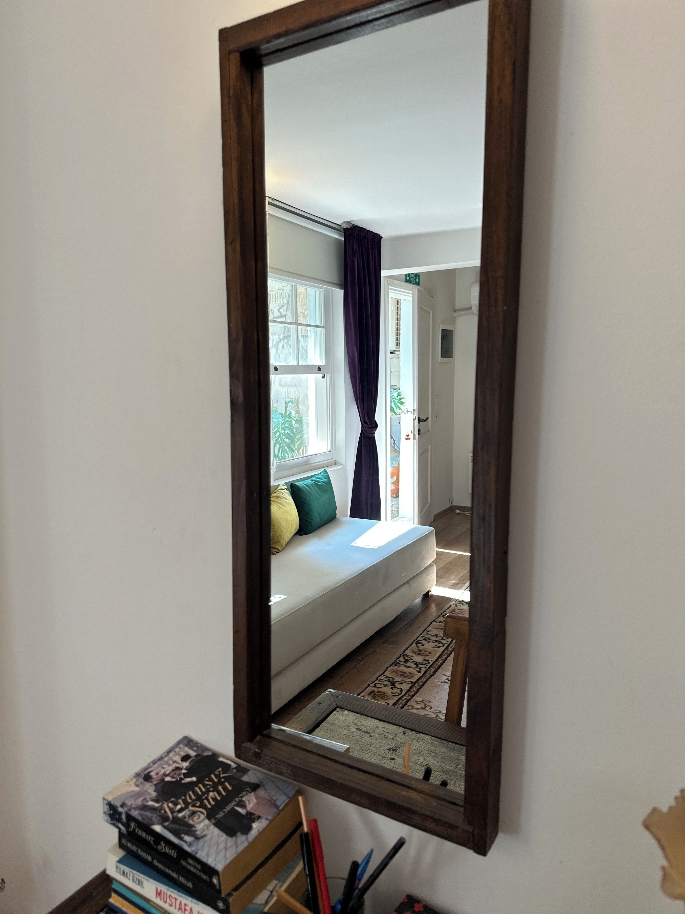 Una habitación con un espejo grande que refleja una cama, una ventana con cortinas y una pequeña mesa con libros y bolígrafos.
